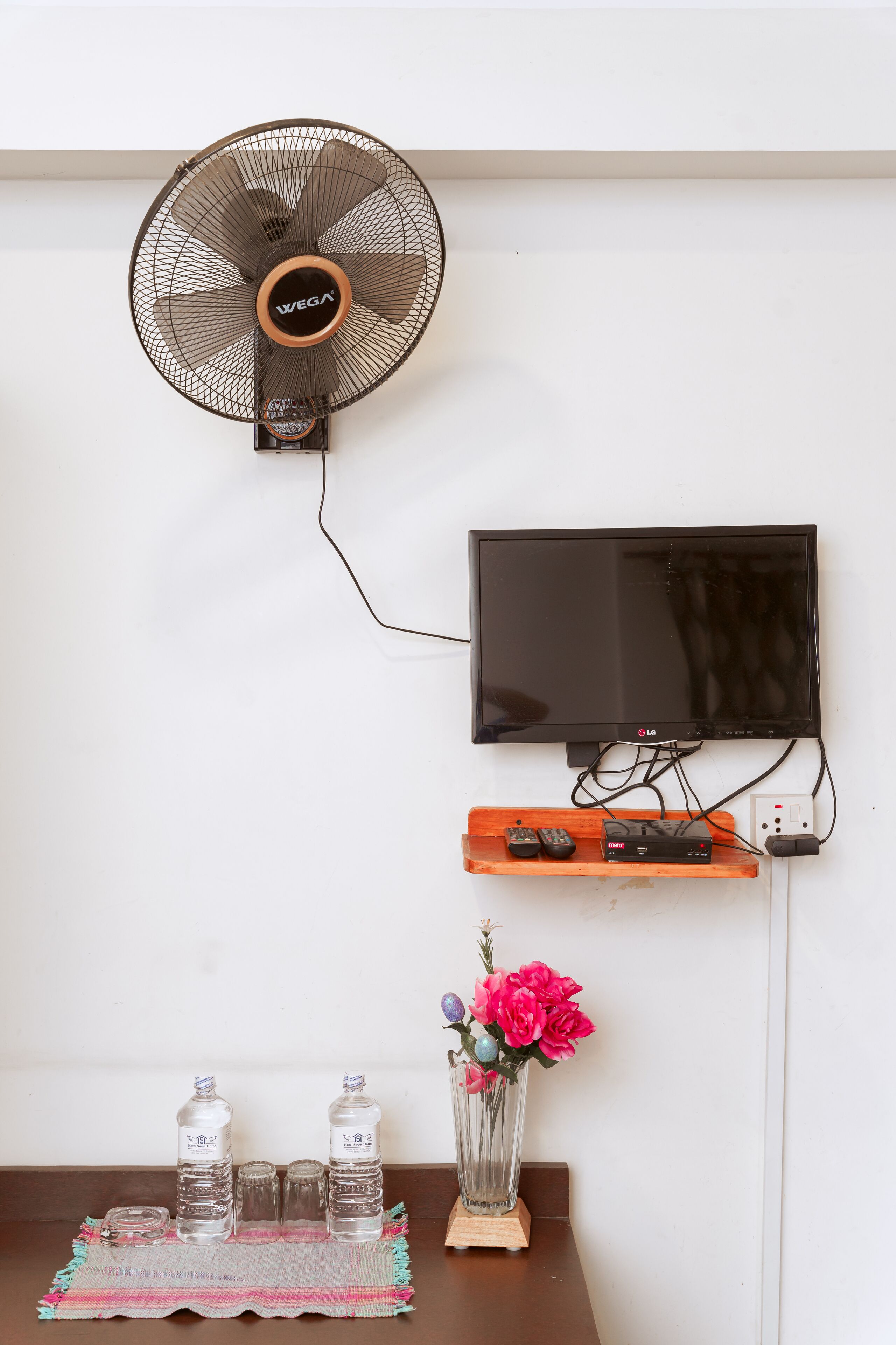 Ein an der Wand befestigter Fernseher mit Fernbedienung, ein Ventilator und eine kleine Ablage mit einer Vase und Flaschen.