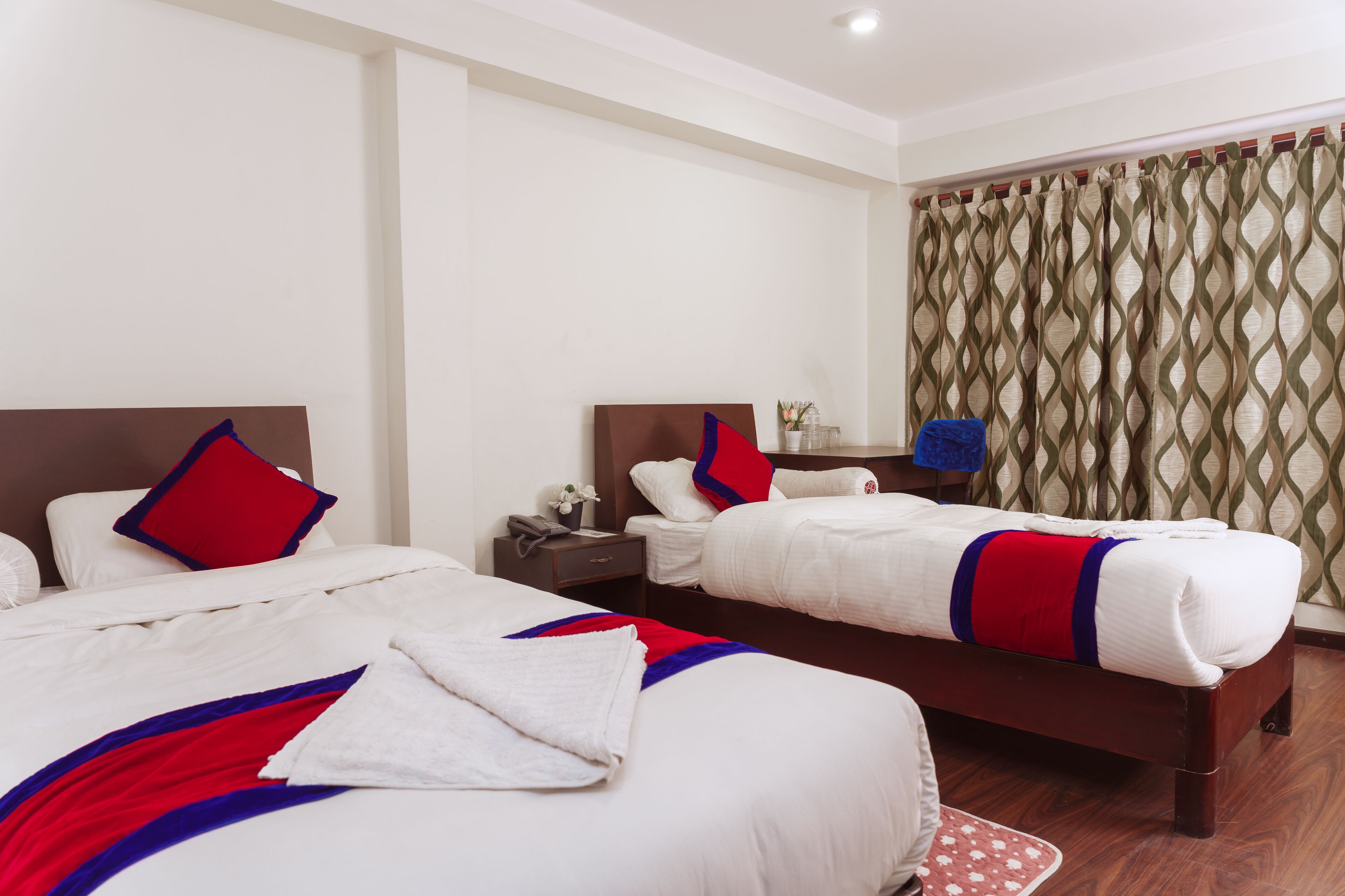 A hotel room with two beds, each with white bedding and red pillows. There is a nightstand with a lamp and a vase of flowers. The room has a wooden floor and a patterned curtain.