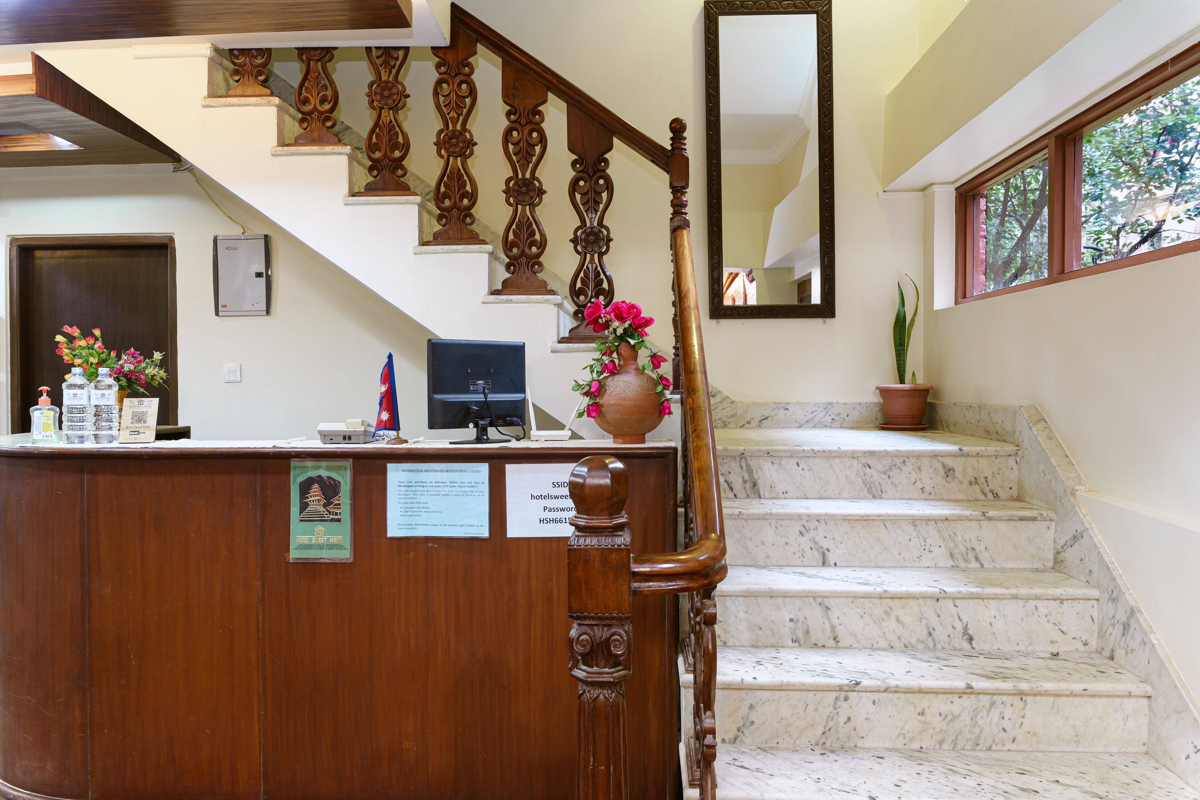 A wooden reception desk with a computer, a vase with flowers, and a sign with room details.