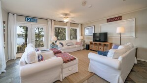 A spacious living room with a large window, a ceiling fan, a television, and a sign indicating 'Arkansas'. - Moonlight Baydream: Lakefront Home w/ Private Dock, Large Patio, Fast WiFi & Near Oaklawn & Downtown (Hot Springs)