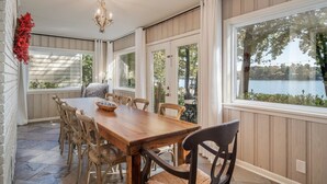 A covered dining area with a wooden table and chairs, large windows with a view of a lake, and a chandelier. - Moonlight Baydream: Lakefront Home w/ Private Dock, Large Patio, Fast WiFi & Near Oaklawn & Downtown (Hot Springs)