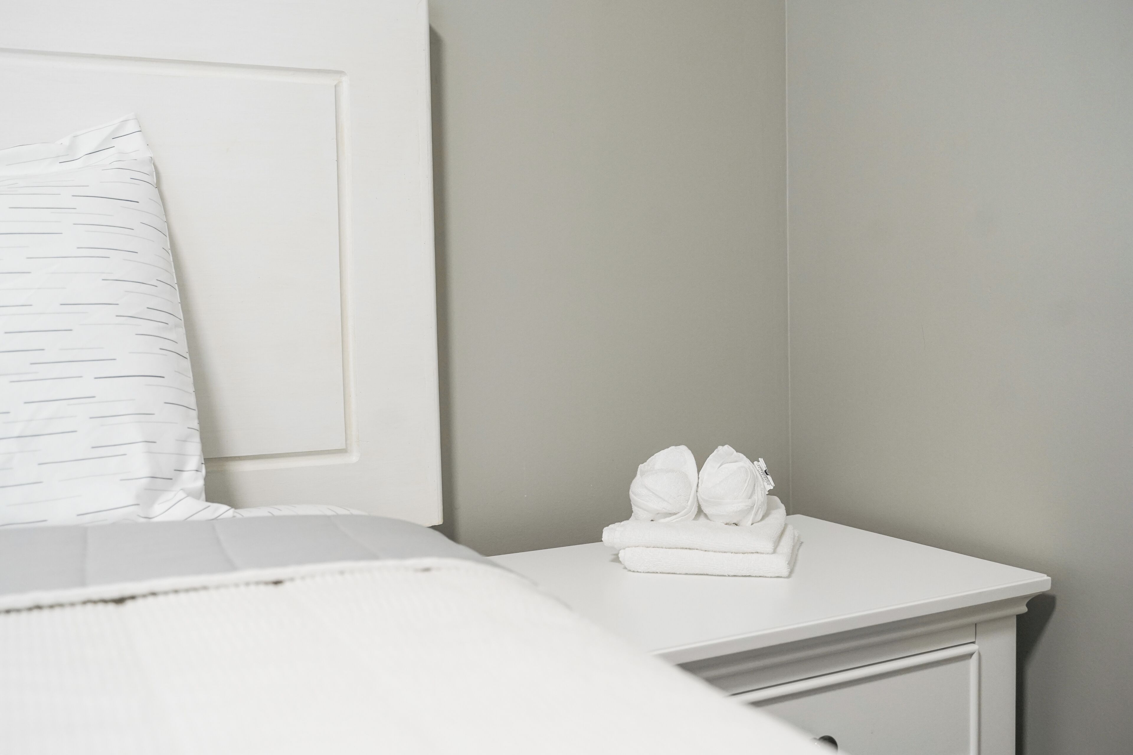 A neatly made bed with a white pillow and a white bedside table with folded towels.