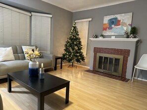 Een woonkamer met een grijze bank, een zwarte salontafel met een bloemenvaas, een kerstboom en een open haard.