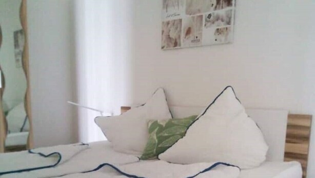 Un lit bien fait, recouvert d’une couverture blanche et bleue, avec une serviette verte et plusieurs oreillers. Une petite table de chevet en bois se trouve à côté du lit.