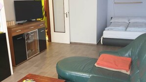 A compact living space with a sofa, a television on a wooden cabinet, a bed with pillows, and a painting on the wall. - FeWo Regenbogen. a colorful start to every day! (Altenau)