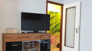 A modern living room with a flat-screen TV on a wooden cabinet, a glass-fronted cabinet with glassware, and a door leading to another room. - FeWo Regenbogen. a colorful start to every day! (Altenau)