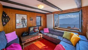 Un salon chaleureux avec des murs en bois, une grande fenêtre donnant sur les montagnes, un canapé agrémenté de coussins colorés et un téléviseur posé sur une table en bois.