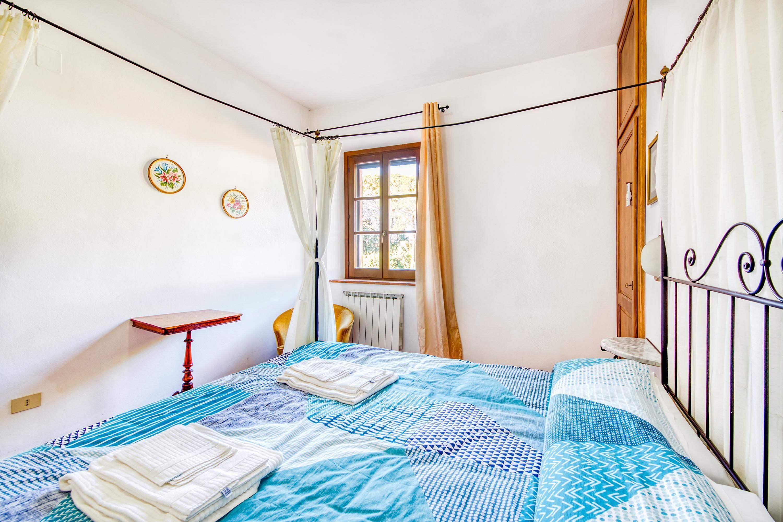 Ein Schlafzimmer mit Bett, Fenster mit Vorhängen, Heizkörper und einem kleinen Tisch.