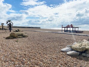 A pebbled beach with a wooden structure and palm trees. - Spacious 2BD Flat With Stunning Views Folkestone (Hythe)