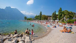 A crowded beach with clear waters, rocky shore, and mountains in the background. - Paulie Apartment in Riva del Garda (Riva del Garda)