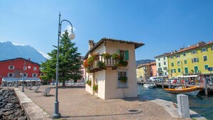 A cobblestone waterfront with colorful buildings, a small kiosk, and a boat docked by the water. - Paulie Apartment in Riva del Garda (Riva del Garda)