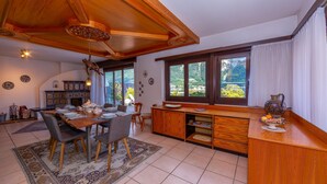 Uma área de refeições com mesa de madeira, cadeiras e tapete. Há também uma cozinha com bancada e armários de madeira. Janelas amplas oferecem vista para o verde lá fora.