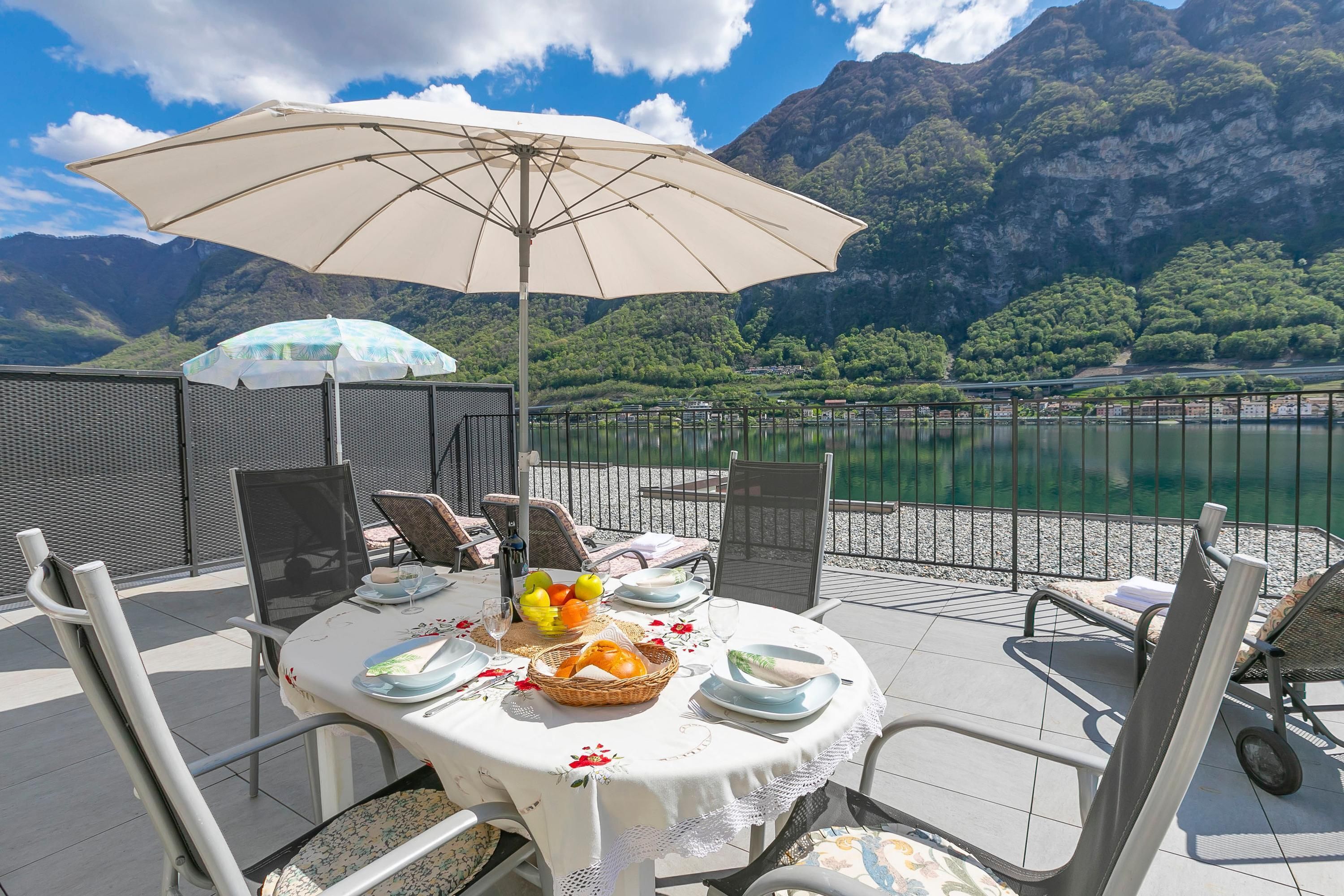 An outdoor dining area with a table set for two, surrounded by chairs, a large umbrella, and a scenic mountain view.