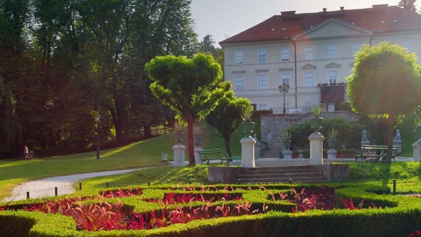 Ein gepflegter formaler Garten mit geschnittenen Hecken, roten Blüten und einem klassizistischen Gebäude im Hintergrund.
