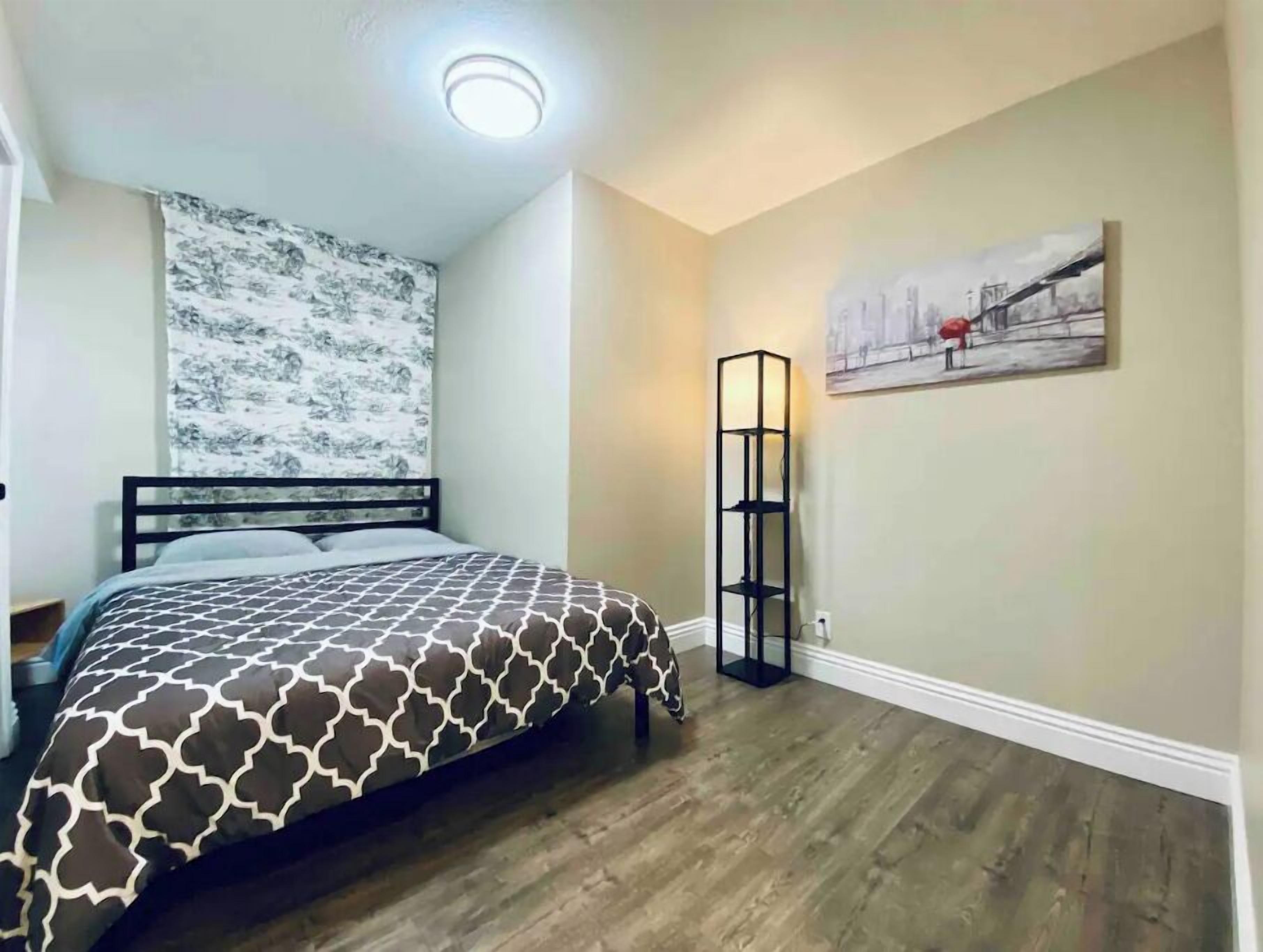 Een slaapkamer met een bed met een patroon, een hoofdbord, een vloerlamp en een stadsgezicht schilderij aan de muur.