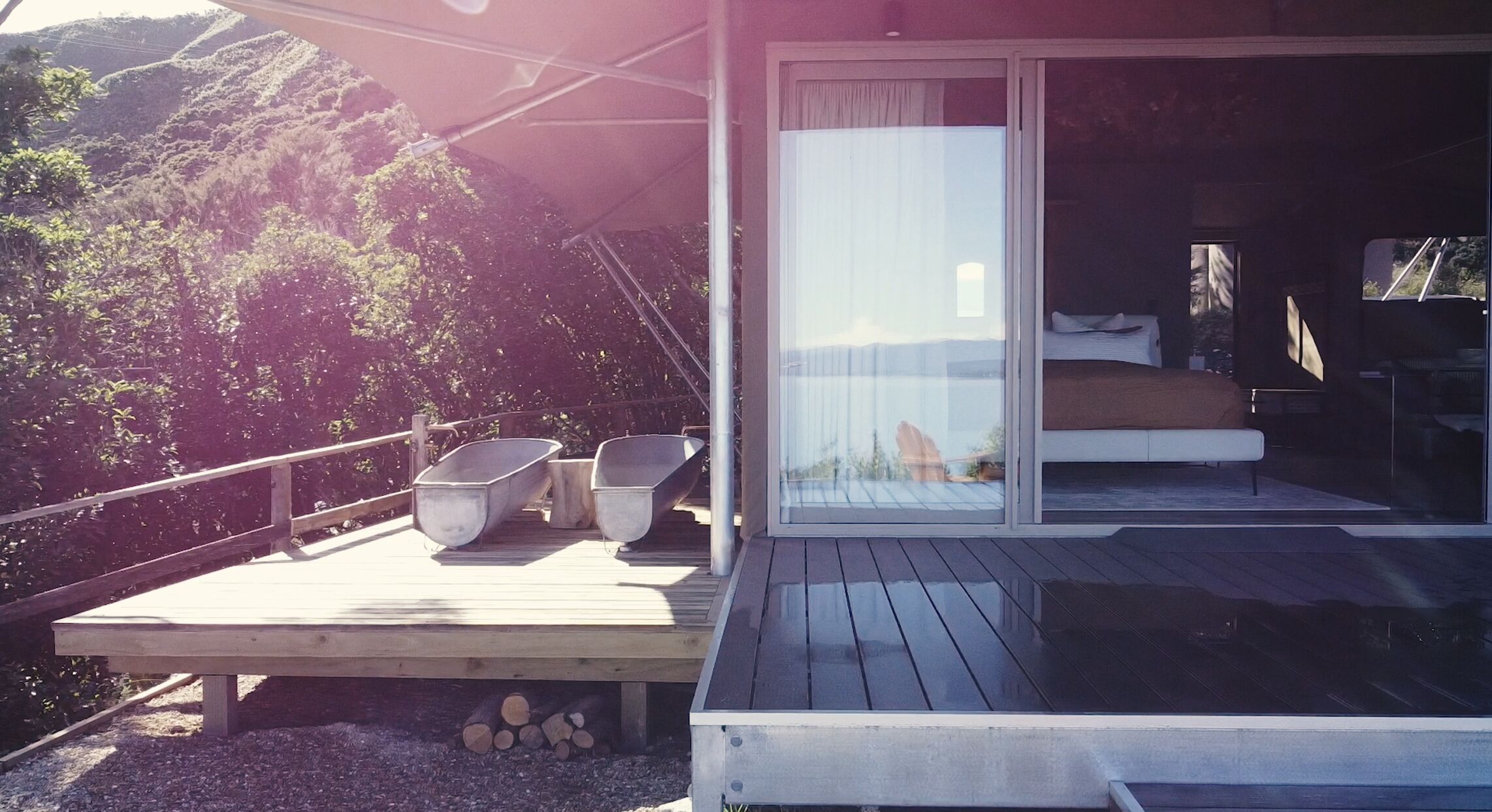 A wooden deck with two metal chairs, a bed with white bedding, and a glass door leading to a bedroom.