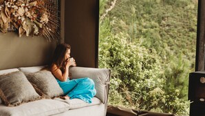 Une personne assise sur un canapé près d’une fenêtre, avec vue sur la verdure.