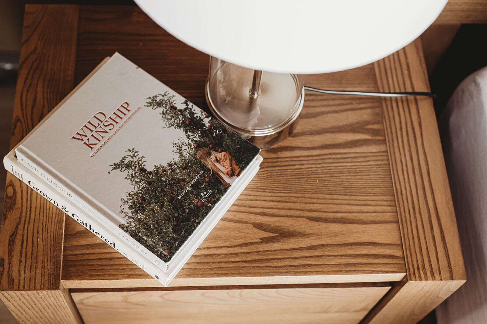 Ein Stapel Bücher mit dem Titel 'Wild Kinship' auf einem hölzernen Nachttisch mit einer weißen Lampe.