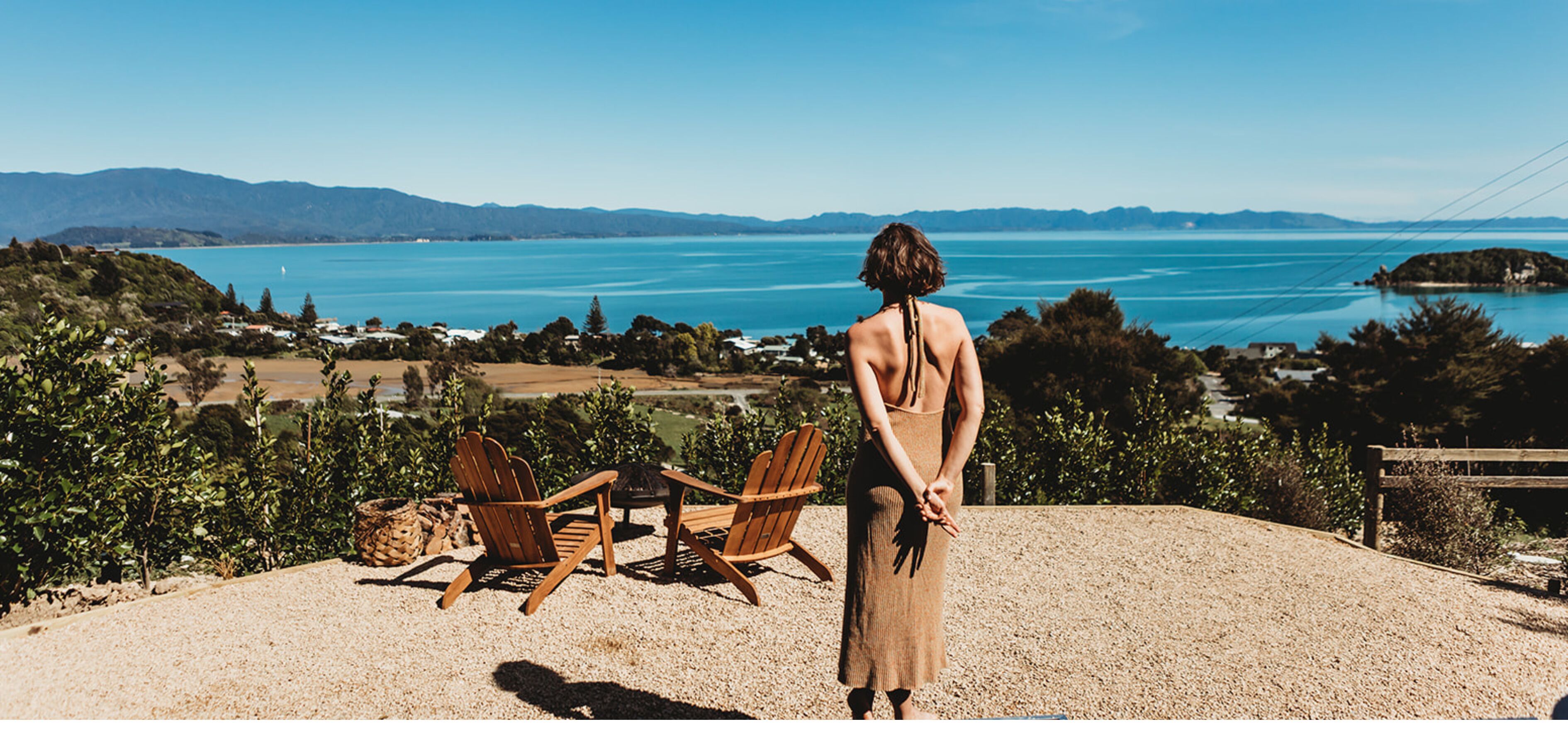 A woman standing on a gravel area overlooking a coastal landscape with a view of the sea and hills.
