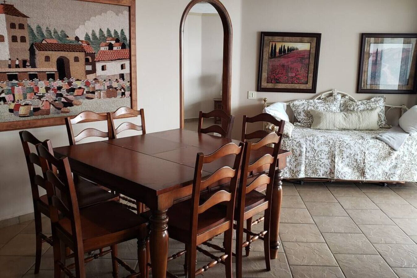 Ein Essbereich mit Holztisch und -stühlen, einem großen Spiegel und einem Bett mit floralem Überwurf.