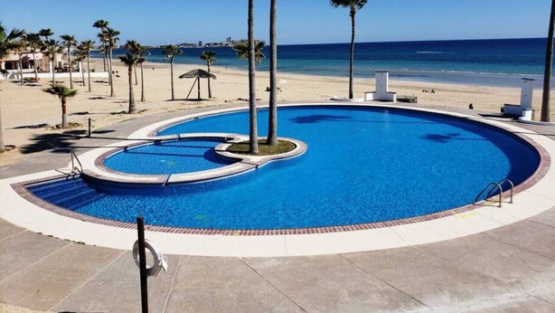 Une piscine circulaire bordée de palmiers, une plage et l’océan en arrière-plan.