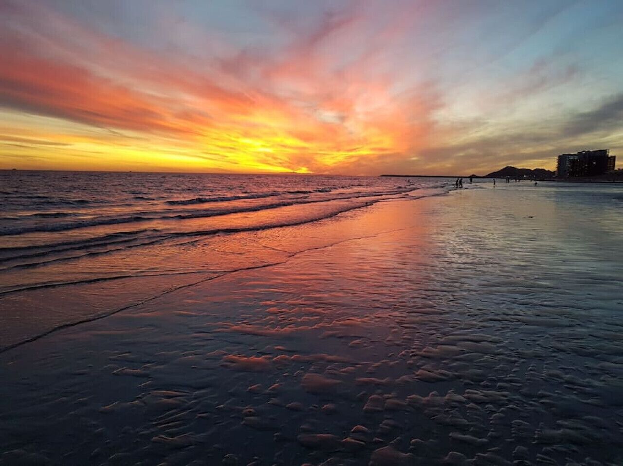Una playa al atardecer con olas, una montaña distante y edificios al fondo.