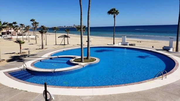 Ein Pool mit gewölbter Kante, umgeben von Palmen und einem Strand im Hintergrund.