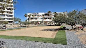 Ein Strandabschnitt mit Sand und einer grünen Rasenfläche, umgeben von einem mehrstöckigen Gebäude mit Balkonen und Palmen.