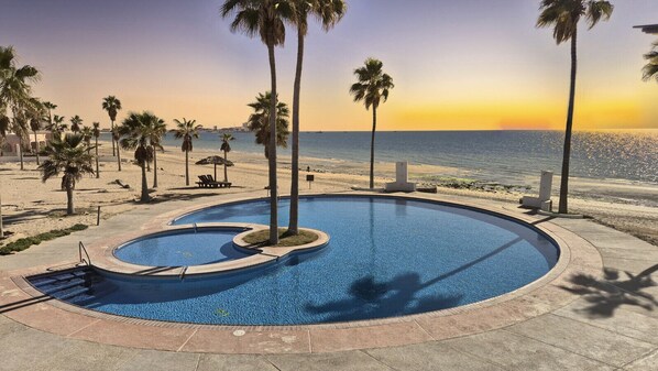 A swimming pool with a curved design, surrounded by palm trees and a view of the ocean at sunset.
