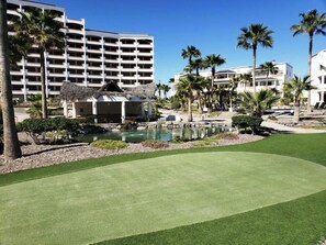 A golf course with a pond, palm trees, and a hotel in the background.