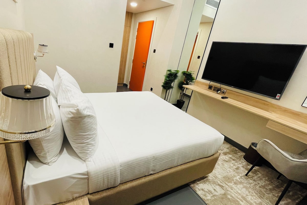 A hotel room with a bed, a bedside lamp, a TV, a chair, and a door.