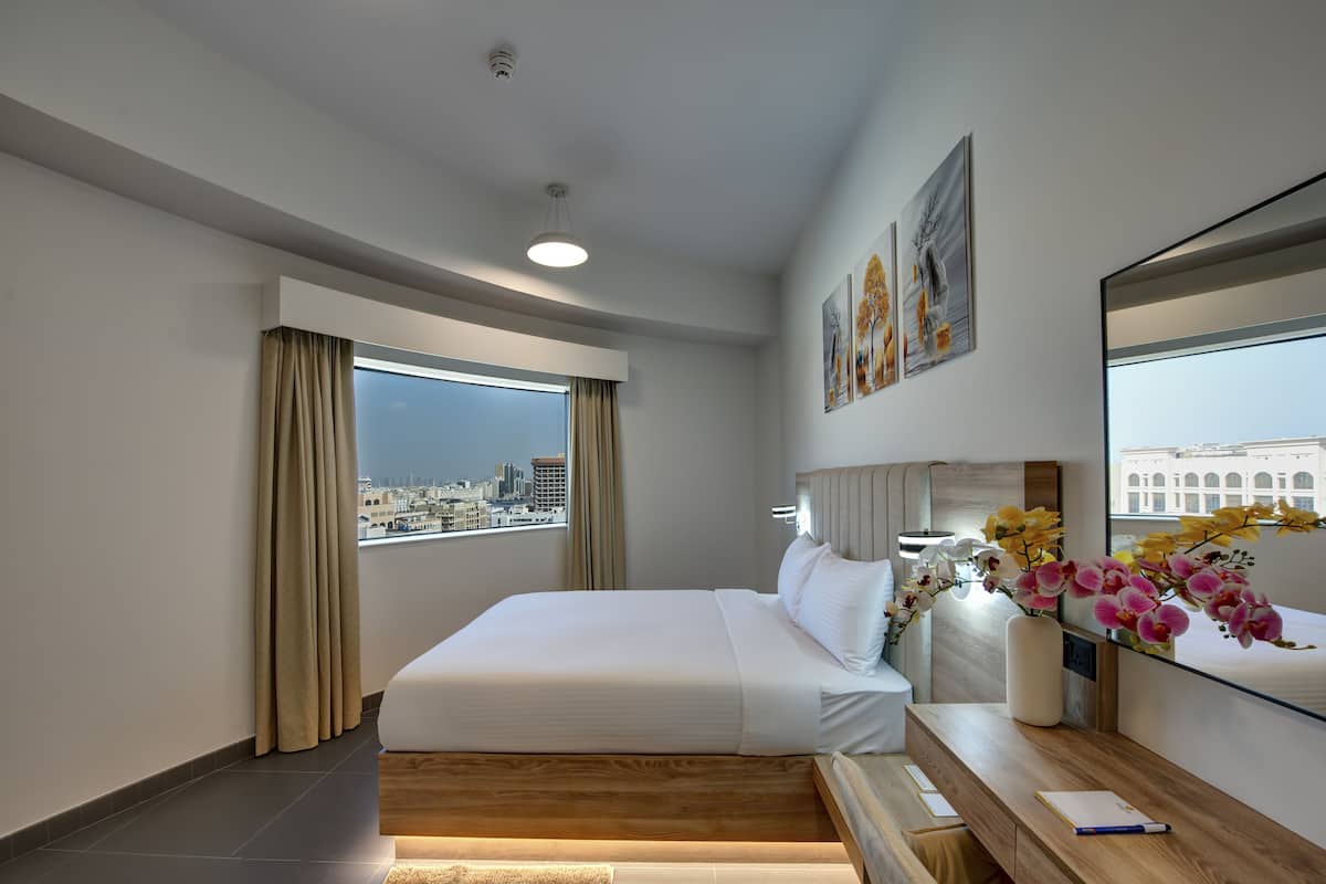 A modern hotel room with a large bed, a desk with a lamp, and a view of the city through large windows.