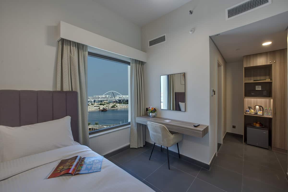 A hotel room with a bed, a desk, a chair, a small kitchenette, and a view of a bridge and water.