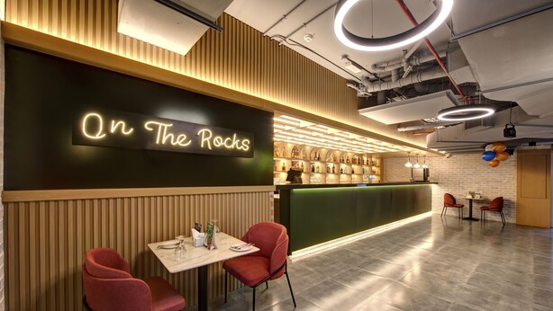 Ein Restaurantinneres mit einem Neonlichtschild mit dem Schriftzug "On The Rocks", Holzvertäfelung, einer Bar mit Flaschen und roten Stühlen.