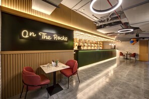 A restaurant interior with a neon sign reading "On The Rocks," wooden paneling, a bar area with bottles, and red chairs. - Wescott Hotel (Dubai)