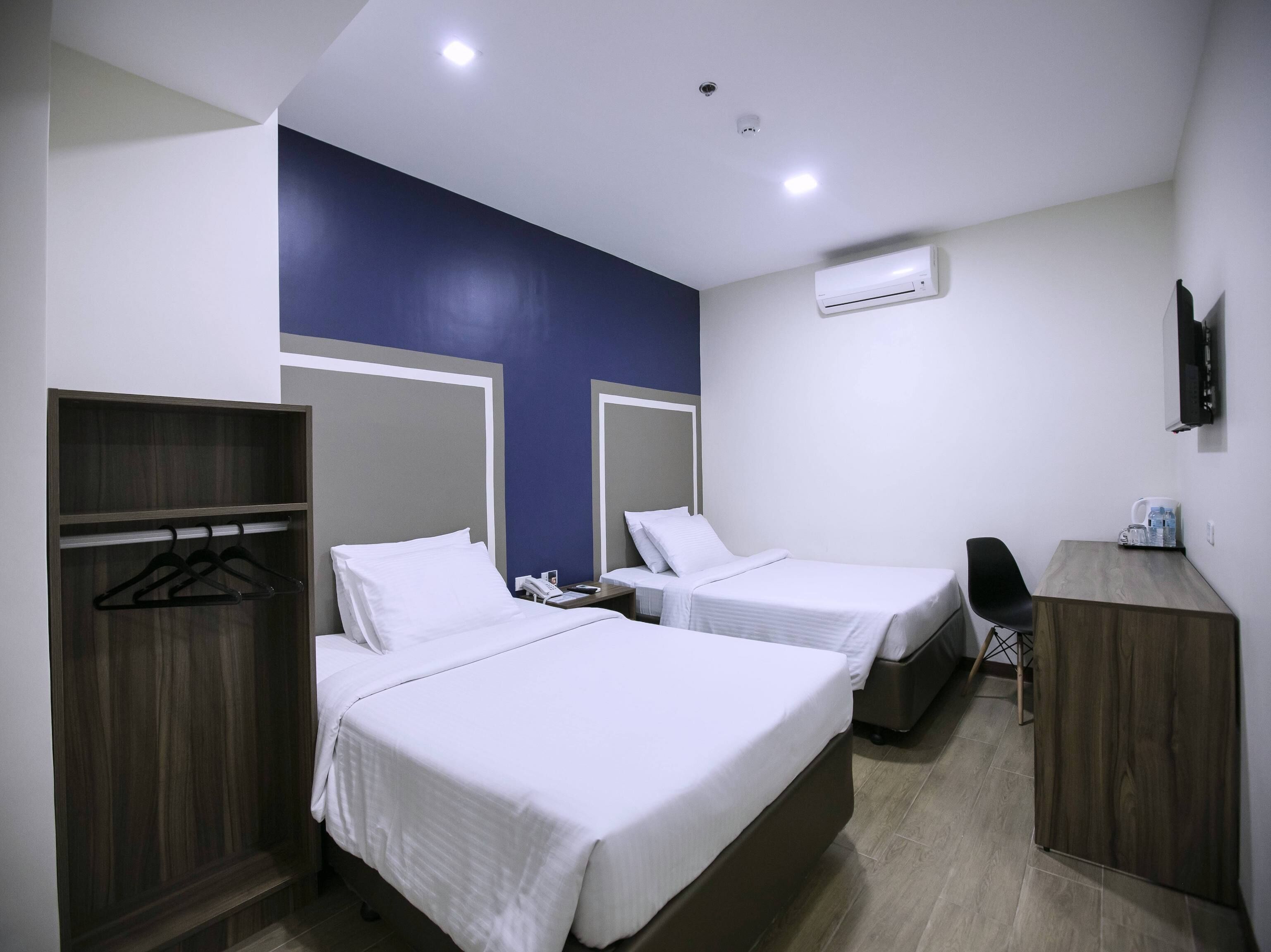 A hotel room with two beds, a wardrobe, a desk, a chair, an air conditioning unit, and a television.