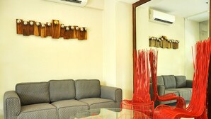 Una sala moderna con un sofá gris, una mesa de centro de vidrio con base roja y una silla roja. Hay decoraciones montadas en la pared y un equipo de aire acondicionado.