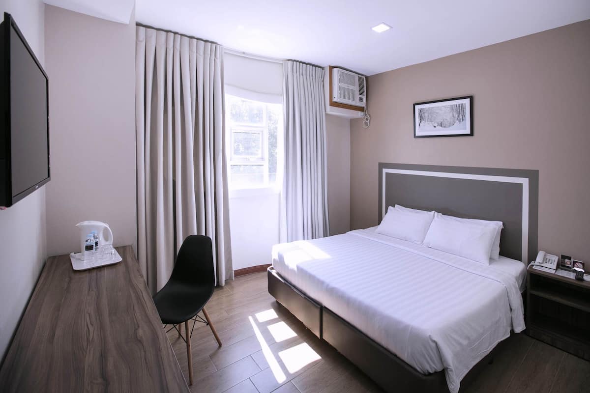 A hotel room with a bed, a desk with a chair, a flat-screen TV, a bedside table with a phone and a lamp, and a window with curtains.