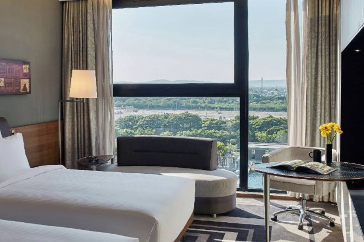 A hotel room with a large bed, a desk, a chair, a lamp, and a view of a cityscape.