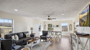 Amplia sala de estar con un sofá gris oscuro, una mesa de centro de madera y un sillón gris. Cuenta con una zona de comedor con mesa y sillas blancas, y un ventanal con vistas al exterior.
