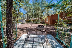 Un banc en métal se trouve sous une structure en treillis verdoyante, dans un espace jardin agrémenté d’arbres, avec un bâtiment en bois en arrière-plan.