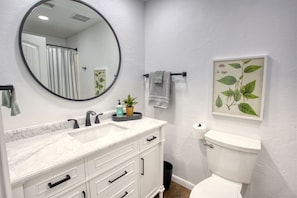 Un baño con un espejo redondo, lavabo blanco y retrete. Hay una planta sobre el lavabo e impresión botánica enmarcadas en la pared.