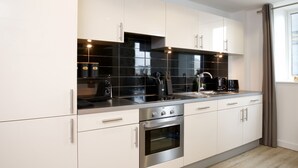 A modern kitchen with white cabinets, black backsplash, stainless steel appliances, and a window with curtains. - Stunning 2 Bed Waterfront Apartment in Media City (Salford)