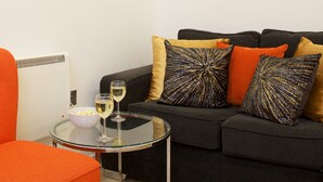 A modern living room with a black sofa, orange armchair, and a glass table with a bowl of popcorn and a glass of wine. - Stunning 2 Bed Waterfront Apartment in Media City (Salford)