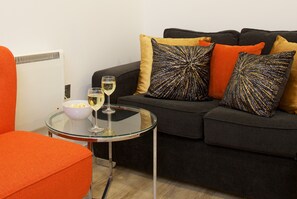 Een moderne woonkamer met een zwarte bank, een oranje fauteuil en een glazen tafel met een kom popcorn en een glas wijn.