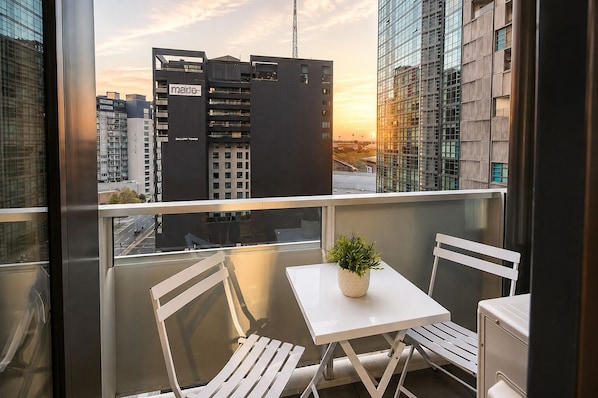 A balcony with a white table and chairs, a potted plant, and a view of a cityscape with modern buildings.