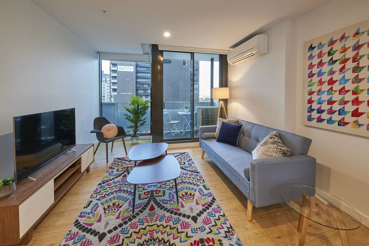 A modern living room with a flat-screen TV, a grey sofa, a colorful rug, and a large window offering a city view.