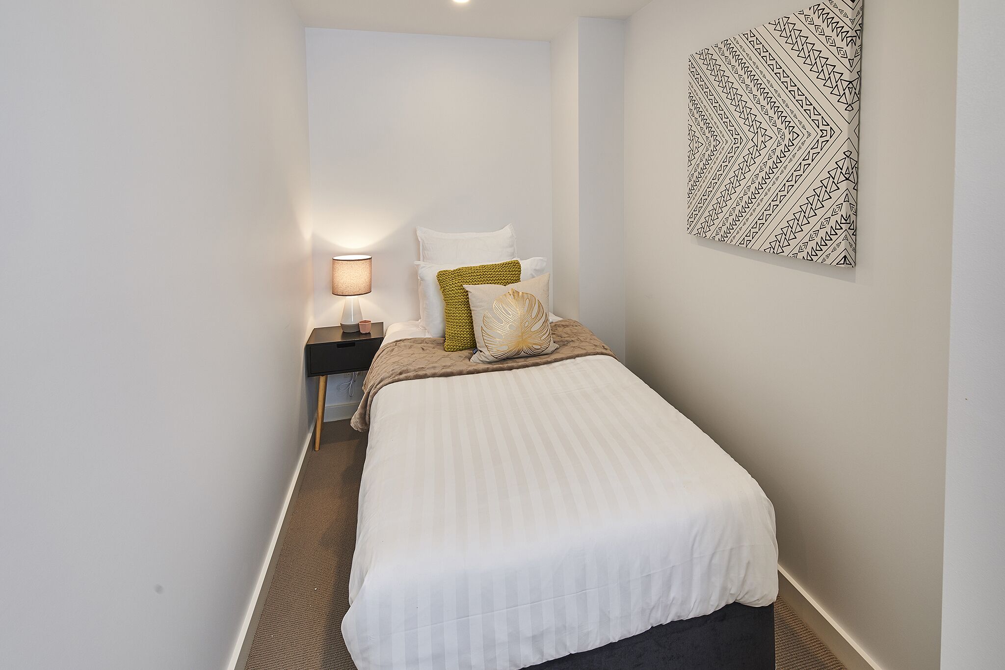 A narrow hotel room with a bed, a nightstand, a lamp, and a wall-mounted artwork.