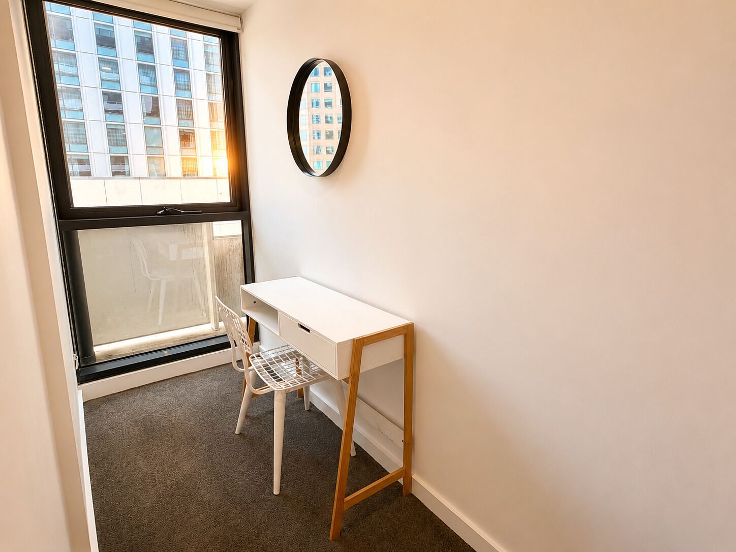 Une petite pièce avec un bureau blanc, une chaise blanche, une fenêtre donnant sur des immeubles et un miroir fixé au mur.