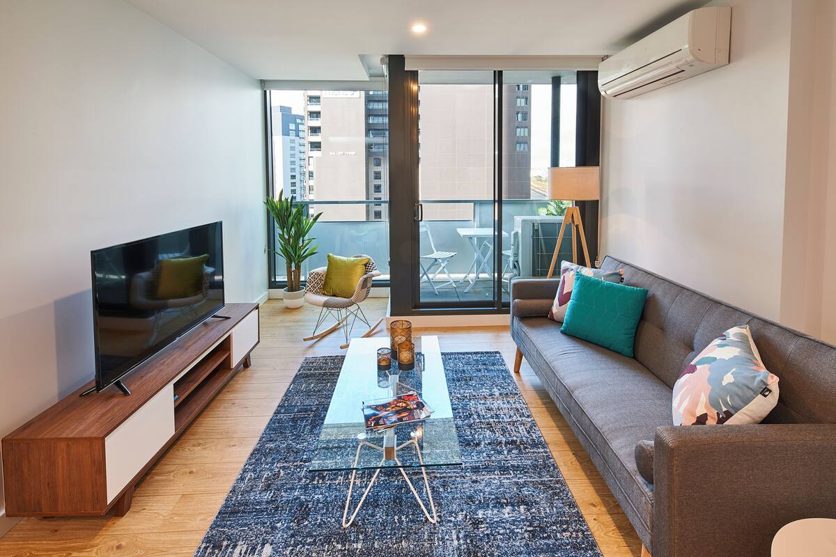 A modern living room with a flat-screen TV, a sofa, a coffee table, and a balcony with a view of the city.